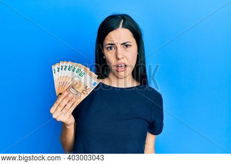 Beautiful young woman holding bunch of 50 euro banknotes in shock face, looking skeptical and sarcastic, surprised with open mouth 