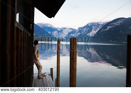 Young Woman Stands By Lake Surrounded By Mountains At Sunset.