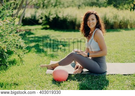 Positive Brunette Woman In Cropped Top And Leggings Poses Bare Feet On Karemat Uses Ball For Gymnast