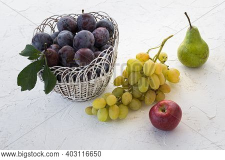 Just Picked Ripe Plums In Wicker Basket And A Bunch Of Ripe White Grapes, An Apple, A Pear On The Wh