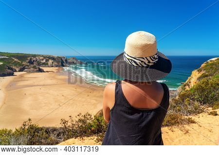 A Woman With A Hat On Her Head, On The Shore, Admires The View Of The Portuguese Beach, Odeceixe.