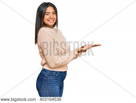 Young latin girl wearing wool winter sweater pointing aside with hands open palms showing copy space, presenting advertisement smiling excited happy 