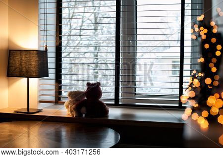 Cozy home evening-two loving embracing teddy bear toys sitting together on wooden windowsill with burning lamp and garland light decor. Living room interior. Filtered horizontal indoor image