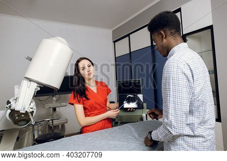 Young Female Caucasian Doctor In Modern Clinic, Holding Tablet Pc And Showing To Her Male African Pa