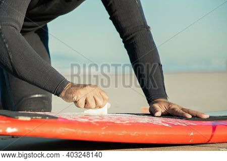 Male Surfer In Wetsuit Waxing Surfboard On Sand On Ocean Beach. Cropped Shot. Surfing And Active Lif
