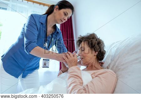 Home Caregiver Helping Elderly Woman Drink Water. Nurse Helping Sick Senior Female Patient Lying In 