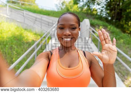 fitness, sport and technology concept - happy smiling young african american woman with wireless earphones listening to music, taking selfie and waving hand outdoors