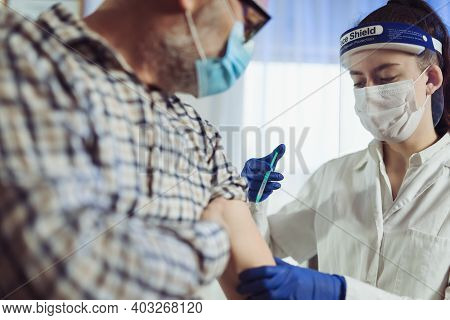 Young Woman Nurse With Surgical Mask And Face Shild Giving Injection To Senior Man. Covid-19 Or Coro