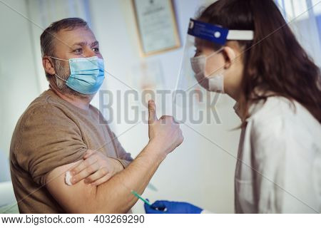 Young Woman Nurse With Surgical Mask And Face Shild Giving Injection To Senior Man. Covid-19 Or Coro