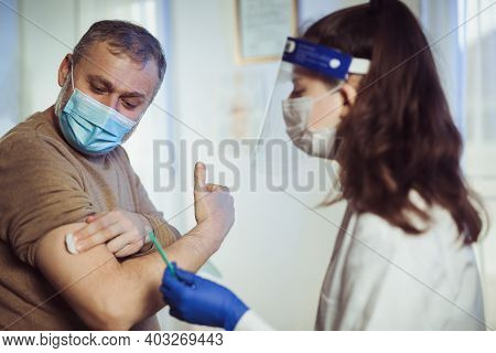 Young Woman Nurse With Surgical Mask And Face Shild Giving Injection To Senior Man. Covid-19 Or Coro