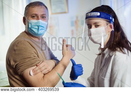 Young Woman Nurse With Surgical Mask And Face Shild Giving Injection To Senior Man. Covid-19 Or Coro