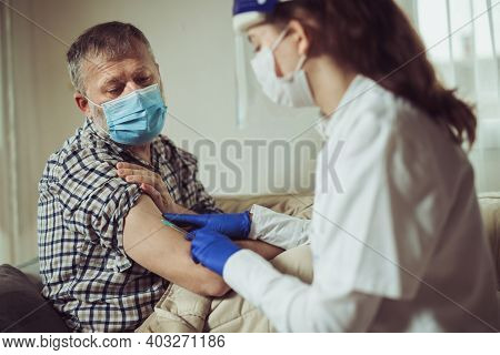 Young Woman Nurse With Surgical Mask And Face Shild Giving Injection To Senior Man At Home Or In A N