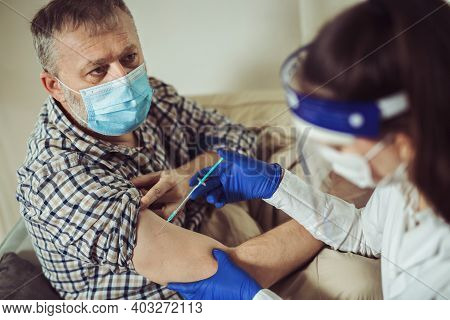 Young Woman Nurse With Surgical Mask And Face Shild Giving Injection To Senior Man At Home Or In A N