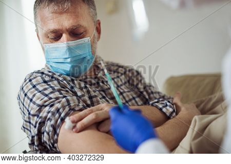 Young Woman Nurse With Surgical Mask And Face Shild Giving Injection To Senior Man At Home Or In A N