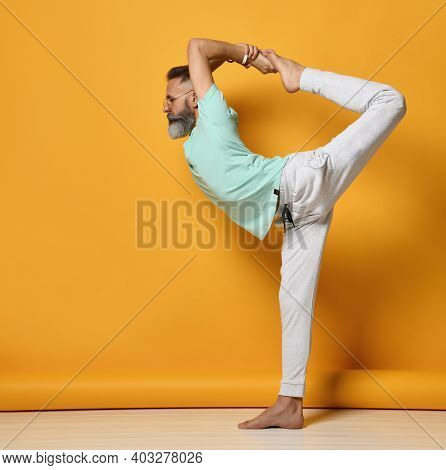 Side View Of A Sporty And Stylish Senior Man Standing On One Leg And Holding The Other Leg, Which Is