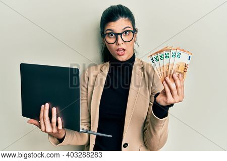 Young hispanic woman wearing business style holding laptop and 50 euros in shock face, looking skeptical and sarcastic, surprised with open mouth 