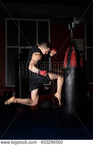 Kickboxing Fighter Performing Jumping Air Kicks With Knee On Punch Bag. Caucasian Man Practicing Mar