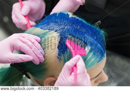 Two Hairdressers Using Pink Brush While Applying Blue Paint To Female Customer With Emerald Hair Col