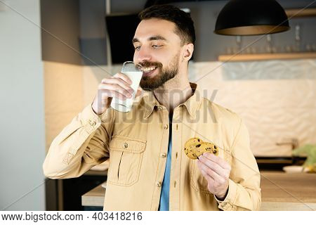 Young Man With Beard And Nice Smile Is Drinking Milk With Cookies In Kitchen In Stead Of Dinner