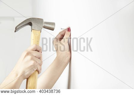 Young Woman With Manicured Hands Nailing A Nail Into A White Wall. Bright Image With Copy Space.