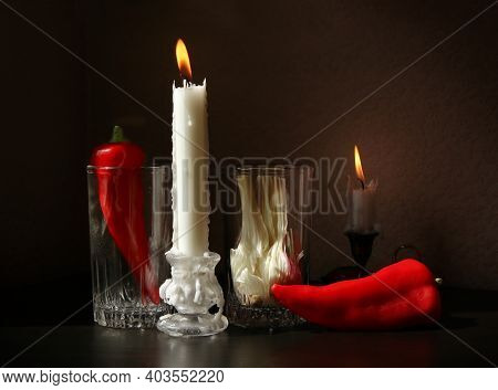 Still Life With Two Candlesticks With Burning Candles, Red Ramiro Peppers And Garlic Bulb In The Gla