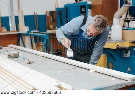 An Elderly Cabinetmaker In Overalls And Glasses Paints A Wooden Board With A Brush On A Workbench In
