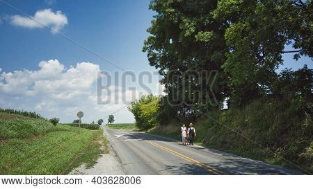 Ronks, Pennsylvania, July 2020 - An Amish Couple One Walking And One On A Scooter On A Road On A Sum