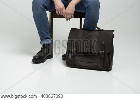 Man In A Blue Jeans And Black Boots Sits On A Chair With A Brown Mens Shoulder Leather Bag For A Doc