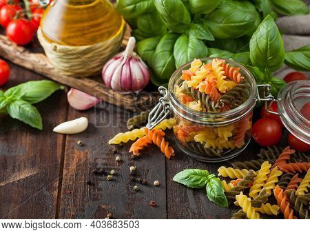 Glass Jar Of Fresh Raw Tricolor Fusilli Pasta With Oil And Garlic, Basil Plant And Tomatoes On Woode