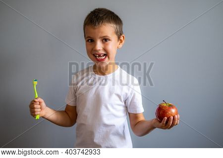 Boy Without Milk Upper Tooth In White T-shirt Holds An Apple And Toothbrush In Hands On The Gray Bac