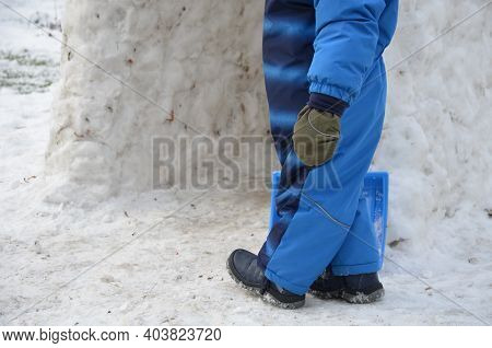 A Small Child In A Winter Jumpsuit Walks With A Blue Plastic Shovel On The Snow. Will Build An Igloo