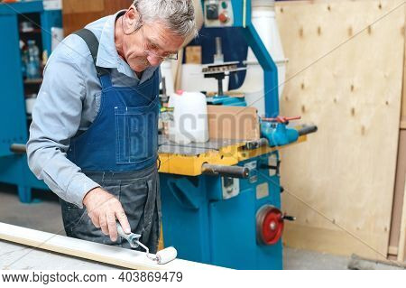 An Elderly Cabinetmaker In Overalls And Glasses Paints A Wooden Board With A Roller On A Workbench I