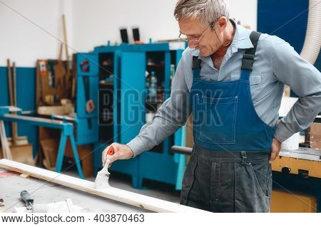 An Elderly Cabinetmaker In Overalls And Glasses Paints A Wooden Board With A Brush On A Workbench In