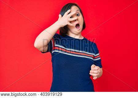Brunette woman with down syndrome wearing casual clothes peeking in shock covering face and eyes with hand, looking through fingers afraid 