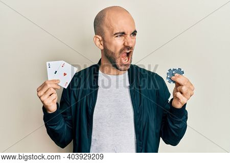 Young hispanic man playing poker holding casino cards and chips angry and mad screaming frustrated and furious, shouting with anger. rage and aggressive concept. 