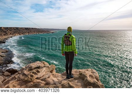 Trailrunner Guy Stands On The Edge Of A Cliff By The Sea On A Sunny Day. View From The Back. Mangyst