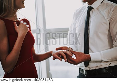 Man Is Proposing To Woman With Engagement Ring Near Window. A Man Puts A Ring On A Woman Who Has Bee