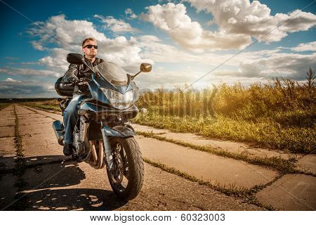 Biker man wearing a leather jacket and sunglasses sitting on his motorcycle looking at the sunset.
