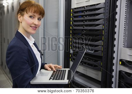 Pretty technician using laptop while working on server smiling at camera in large data center