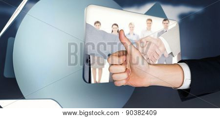 Hand showing thumbs up against screen displaying handshake and business people in digital inter