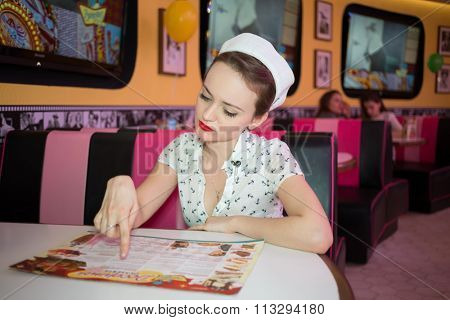 Concentrated young woman in retro dress sitting in bar and looking to menu.