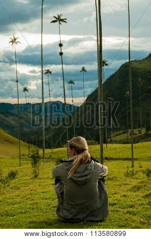 Girl with grey jacket sitting on green grass watching tall palm trees in Cocora Valley