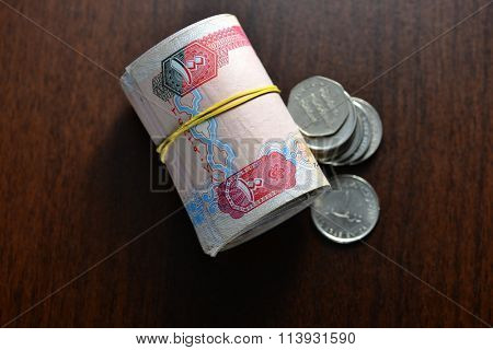 Hundred dirham currency notes rolled and tied with rubber band. UAE currency notes and coins placed on table. View from top.