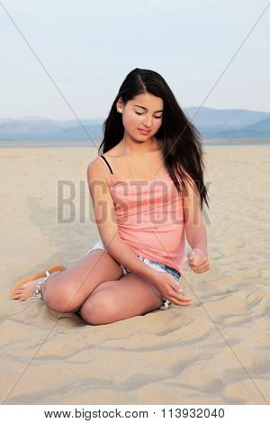 Young european teenage girl with long dark hair sitting and playing with sand on a beach.