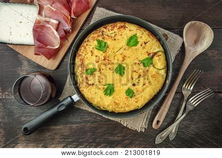 An overhead photo of a Spanish tortilla with potatoes and zucchinis in a tortillera, with jamon, cheese, a glass of red wine, a wooden ladle, and two forks, with a place for text