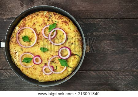 An overhead photo of a Spanish tortilla with potatoes and zucchinis in a tortillera, on a dark rustic wooden background with a place for text
