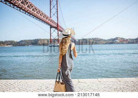 Lifestyle portrait of a woman walking on the riverside with beautiful iron bridge on the background in Lisbon city, Porugal