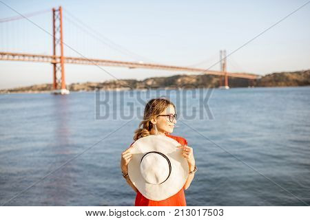 Portrait of a woman in red dress with sun hat standing on the riverside with beautiful iron bridge on the background in Lisbon city, Porugal
