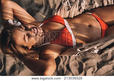 Carefree and happy. Top view of attractive young woman in swimwear smiling and keeping hands behind head while lying down in hammock on the beach