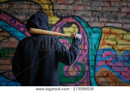 A street hooligan in a black hood on a construction site stands with a wooden bat on his shoulder against the backdrop of a brick wall with a graphite.
